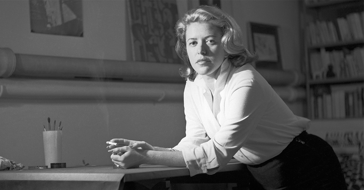 Imagem em preto e branco de Hilda Hilst em pé, com os cotovelos apoiados na mesa e segurando um cigarro na mão direita